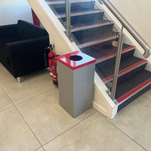 office recycling bin with silver body and red top at bottom of stairs